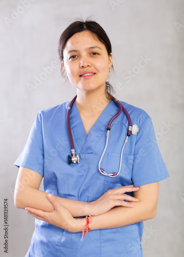 Friendly young female professional doctor in blue scrub with phonendoscope around neck posing against gray studio background, looking at camera with smile..