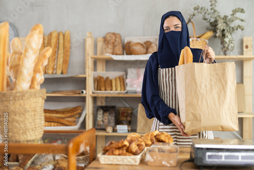Obraz na płótnie Female vendor in traditional Muslim clothing, niqab, holds out a paper bag with fresh bread at a bakery w salonie