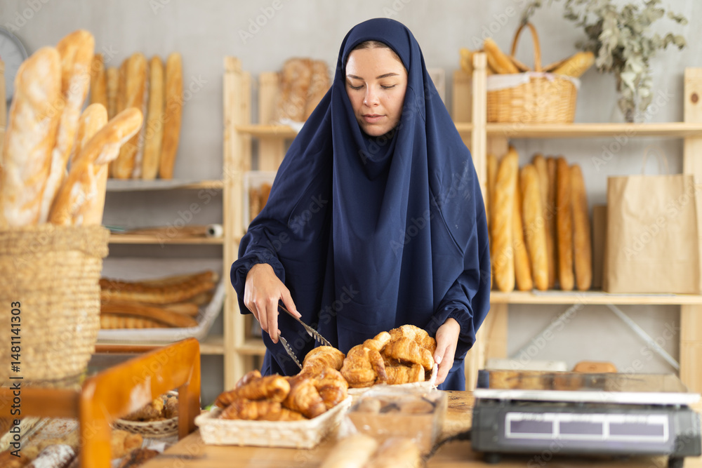Girl in traditional Muslim clothing selects and places fresh hot croissants in a plastic bag