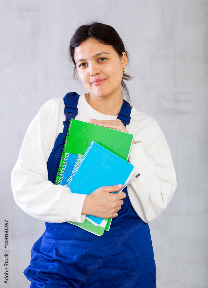Portrait of young female mechanic in blue overalls holding workbooks with orders, posing in studio against gray plain background, looking confident and positive. Professional activity concept..