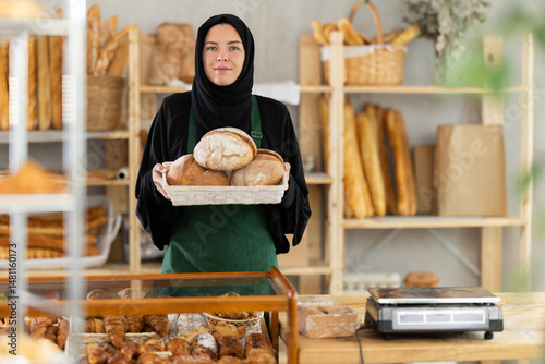 Muslim European girl works in sales area of bakery, selling bread in paper bag. She holds basket filled with fresh fragrant loaves of bread. Store employee near counter sells bakery products