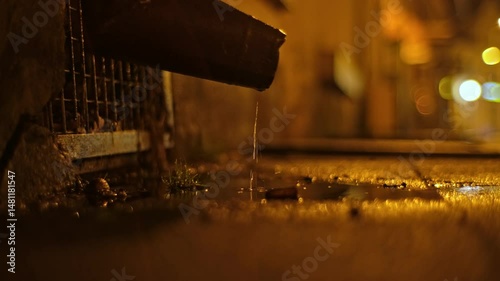 Rainwater Dripping from Metal Gutter Chute into Puddle on Pedestrian Sidewalk in Gloomy City at Night Scene