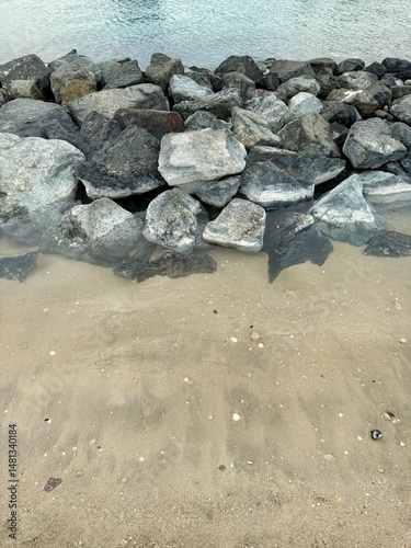 Large gray rocks piled along a sandy beach with scattered seashells, partially submerged in clear water with ripples near the shoreline.