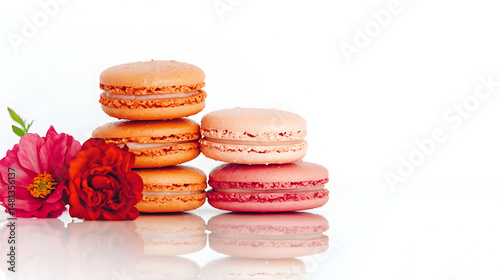 Stacked orange and pink macarons beside red and pink flowers, placed on a glossy surface with wide white space on the right, perfect for marketing text or branding.