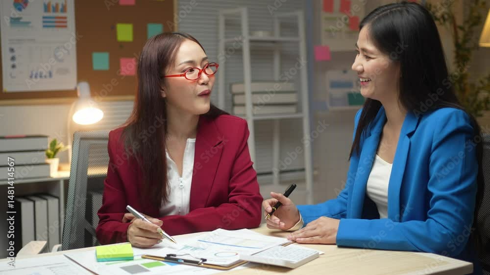 Two women are sitting at a desk, talking and writing. One of them is wearing a red jacket Two women are sitting at a desk, talking and writing. One of them is wearing a red jacket