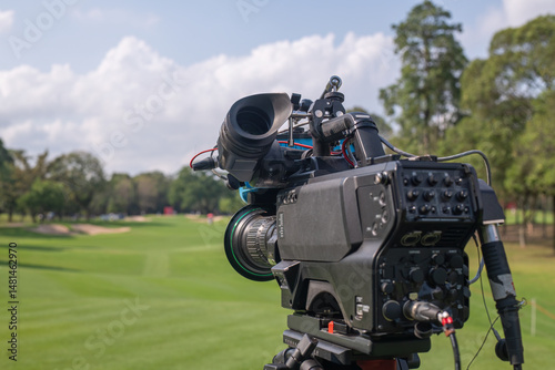 Professional broadcast video camera set up on a tripod overlooking a golf course. The camera is focused on a lush green fairway, likely capturing footage for a live or recorded golf tournament.