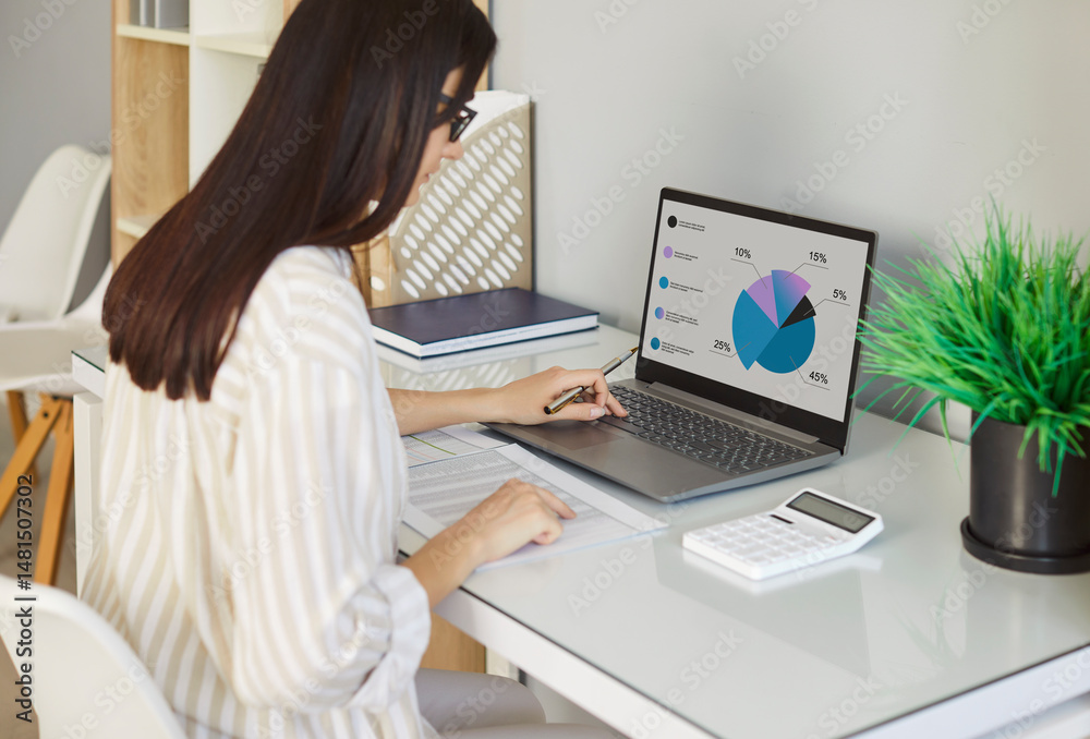 Fototapeta premium Portrait of a concentrated young business woman accountant sitting at the desk and working on a laptop computer at office or at home and looking to the monitor screen. Accounting job and finance