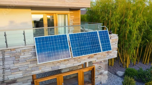 Modern house with stone facade and solar panels mounted on balcony railing, surrounded by greenery, representing eco-friendly living, renewable energy power, sustainable residential design, sunlight.