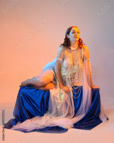 Fantasy studio portrait of a red-haired mermaid faery model wearing a white pearl gown, fairytale costume. posing in a classical sitting position with flowing fabric, ethereal golden studio lighting