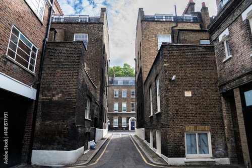 Fototapeta Naklejka Na Ścianę i Meble -  Narrow Alley Between Classic Brick Buildings Under Clear Sky, Featuring Historic Architecture and Small Residential Windows in a Peaceful Urban Setting.