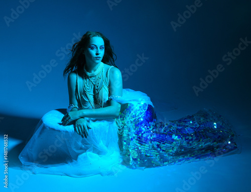 Fantasy studio portrait of a red-haired mermaid faery model wearing white pearl gown, fairytale costume. classical sitting pose, with flowing fabric, colourful neon moody lighting on studio background