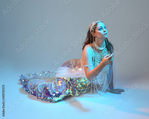 Fantasy studio portrait of a red-haired mermaid faery model wearing white pearl gown, fairytale costume. classical sitting pose,, ethereal moody blue lighting as if underwater on studio background