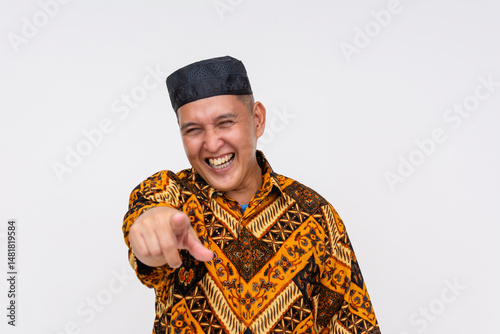 Middle-aged Indonesian man bursting into laughter while pointing directly ahead, dressed in Batik shirt and traditional songkok. Isolated on a light background.