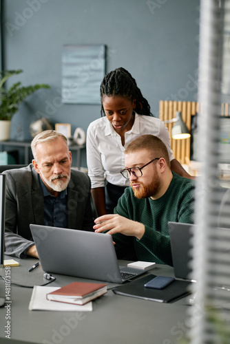 Multiracial business team comprising two men and a woman collaborating on a project in modern office setting, using laptops and other technology devices while discussing work