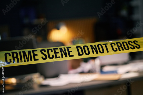 Yellow crime scene tape cordoning off an area inside building with various blurred objects in background. Focus is on crime scene tape, creating suspenseful atmosphere