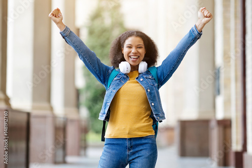African American female student raising her arms in celebration, passed exam successfully, joyful black woman wearing denim jacket and headphones, smiling brightly on university campus, copy space