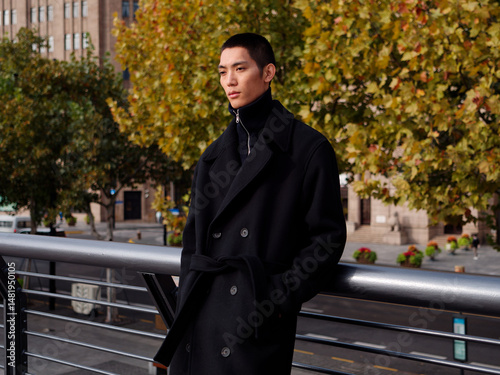 Portrait of handsome Chinese young man wearing black overcoat posing in the street, young guy with black short hair all in black with urban background. Male fashion, cool Asian young man lifestyle.