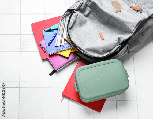 An open backpack spilling out colorful notebooks, pens, and a folded lunchbox resting on white tiles.