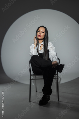 Stylish woman in black and white outfit posing on a chair in studio with dramatic lighting. Confident fashion look, long dark hair, bold makeup, minimalist and edgy atmosphere.