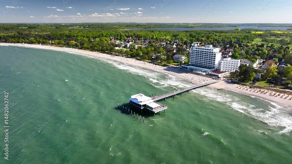 Aerial footage of Baltic Sea beach resort Timmendorfer Strand with pier stretching into green sea, beachfront hotel, white beach chairs and coastal town framed by public health resort park.
