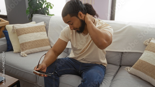 Young man with a beard feeling neck pain while sitting on a sofa in a modern living room, holding glasses in one hand and appearing to be in discomfort.
