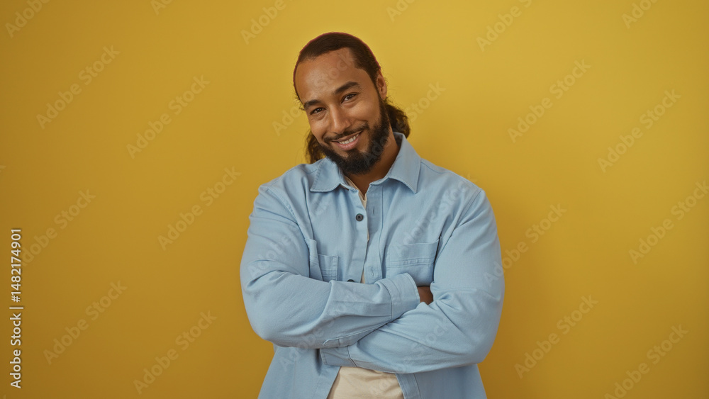 Smiling man with beard stands against bright yellow background wearing a light blue shirt, expressing confidence and positivity with arms crossed and a welcoming demeanor Smiling man with beard stands against bright yellow background wearing a light blue shirt, expressing confidence and positivity with arms crossed and a welcoming demeanor