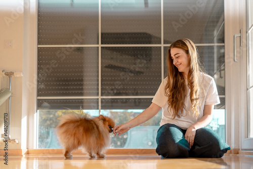 Happy Young Woman Sitting on Floor with Her Pomeranian Dog at Home by the Window photo