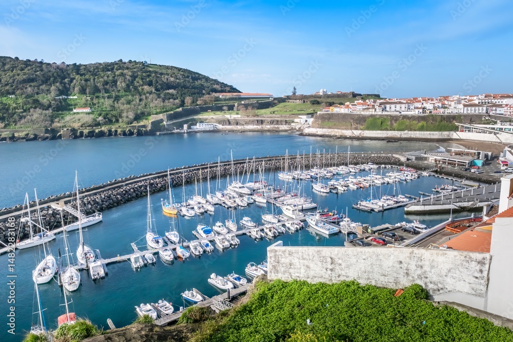 Fototapeta premium Scenic aerial view of marina with sailboats and yachts in natural harbor. Angra do Heroismo marina, historic fortress on Monte Brasil green hillside under blue sky, Terceira Island, Azores, Portugal.