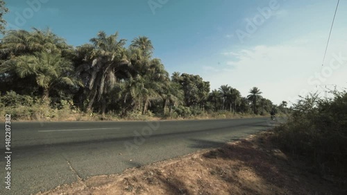 Motorbike entering frame on sunny Casamance road at midday, surrounded by green bush and tropical vegetation, filmed in rural Senegal under clear blue sky and strong African sun