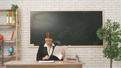 Young woman college teacher at desk in classroom in front of chalkboard reading grading homework or tests of students. Education concept.