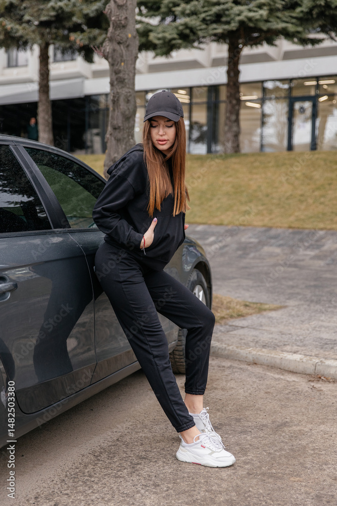 Fototapeta premium Cute girl model in a sporty comfortable casual black outfit with a hoodie and sweatpants posing shows a quality ot clothing