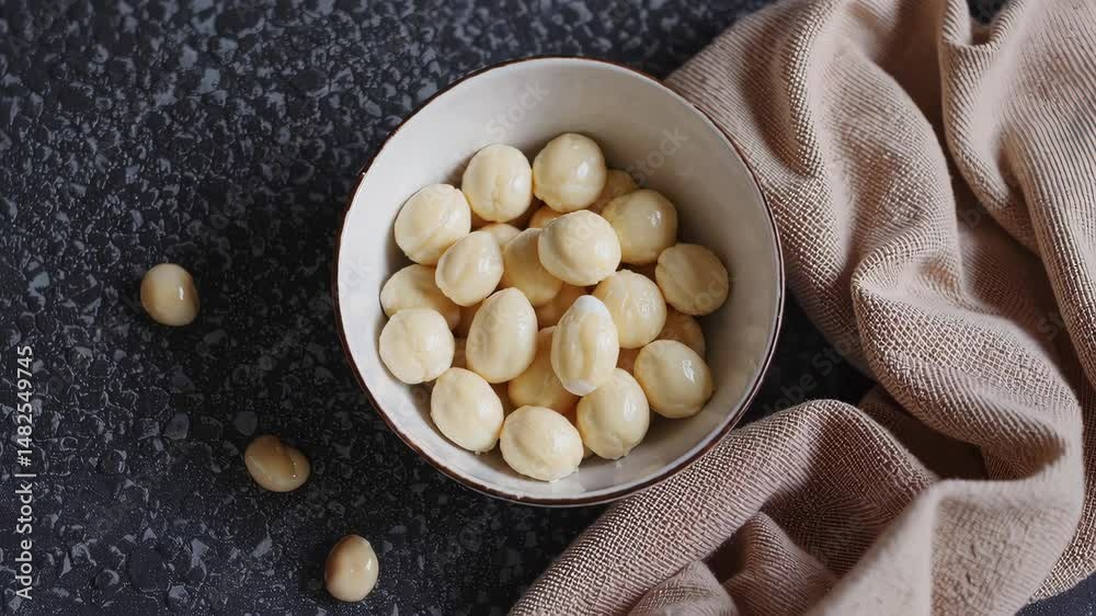 Bowl of shelled macadamia nuts on black textured surface with beige cloth, natural food product scene in minimalistic modern style loop
