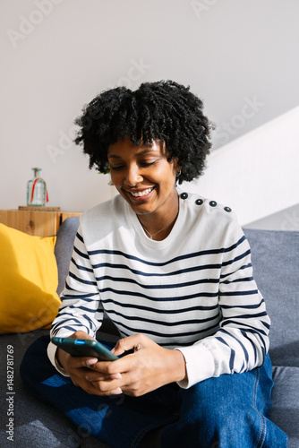 Vertical shot of happy young Latin American girl using mobile phone relaxing on sofa at home, enjoying social media content or watching funny videos