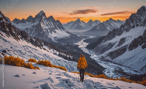 Man in a yellow jacket is walking on a snowy mountain trail. The mountains in the background are covered in snow and the sky is a beautiful orange and pink color. Concept of adventure and exploration