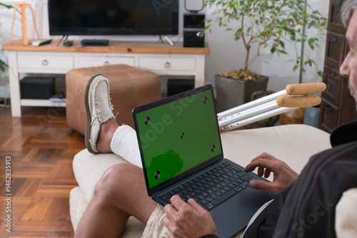 Man with leg in plaster cast using laptop with green screen at home