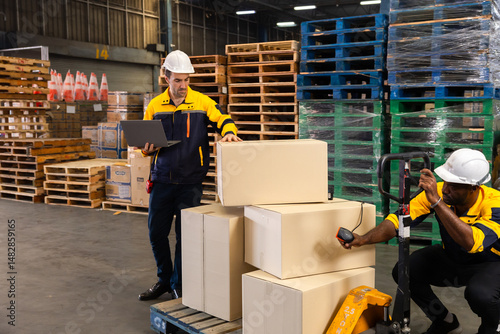 Warehouse team collaborates as one worker crouches to scan boxes with handheld scanner while colleague updates data on laptop, ensuring cargo tracking in industrial aisle. Diverse males, mid-30s