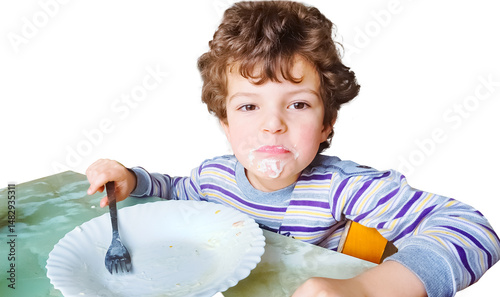 A little boy with a dirty mouth looks slyly at the camera, an empty plate on the table in front of him. Childish facial expressions, mischief and appetite. Isolate