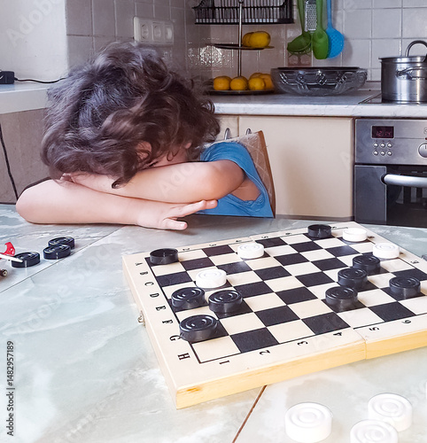 Portrait of little boy playing checkers: he head on hands, tears on face, indoor game scene, emotions of disappointment and frustration