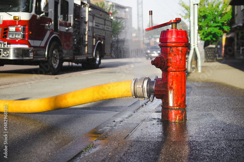 Close up of fire hydrant with hose attached and defocused fire truck. Supply line or hydrant line width large diameter. Water spaying from hole. Fire hall responding to fire in city. Selective focus.