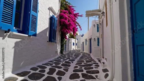 Mykonos, Greece, is depicted in a traditional whitewashed street of Mykonos town featuring blue windows and doors on a sunny summer morning with an empty alleyway at sunrise