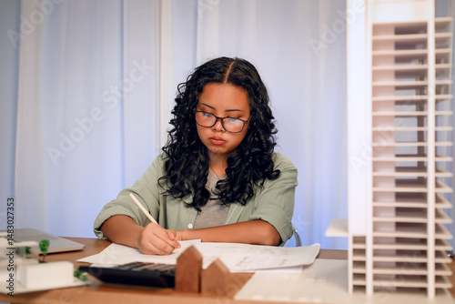 University students young woman with glasses focuses on writing or sketching at her desk, surrounded by architectural models and design tools, representing creativity and concentration in project.