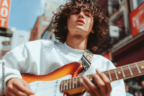 Young musician wearing a white shirt is playing an electric guitar outdoors on a sunny day. He is standing in a city street with his eyes closed