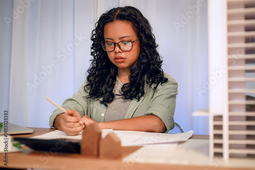 University students young woman with glasses focuses on writing or sketching at her desk, surrounded by architectural models and design tools, representing creativity and concentration in project.