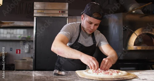 Pizzaiola spreading filling at dough. Male working at cafe as pizzaiola. Man cooking pizza at background of oven. Male wearing apron and cap at kitchen. Pizza baker in professional uniform.