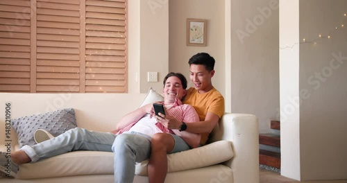 Diverse male couple on couch joyfully sharing engagement news with smartphone, showing ring, at home