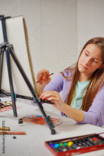 Vertical close-up of a little girl with long brown hair sitting at a desk with an easel