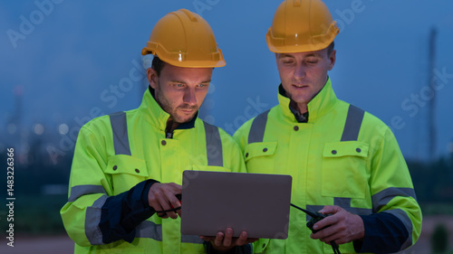 caucasian white engineers in safety uniforms and helmets using laptop and radio for site communication. Concept of teamwork, inspection, and operations at an industrial facility.Construction of Oil, 