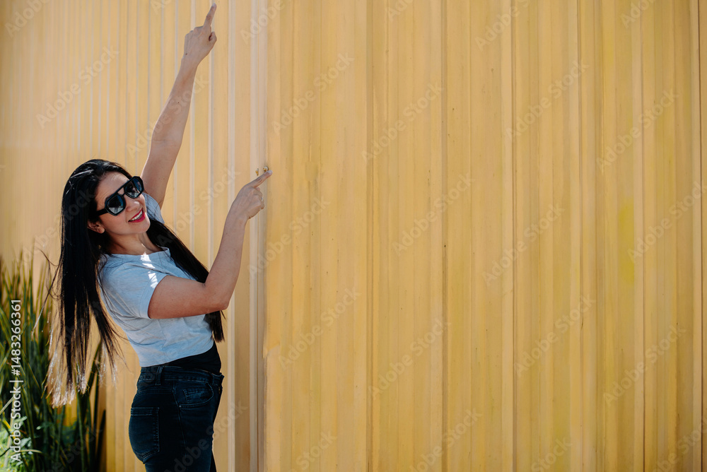 Fototapeta premium Trendy woman wearing sunglasses, smiling and pointing upward, yellow background enhances positivity.