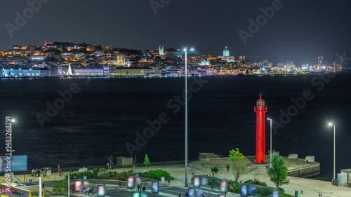 Aerial night timelapse of Cacilhas bus and railway station with the Tagus River, Lisbon glowing skyline and a red lighthouse. Boats passing by illuminate the water. Portugal.