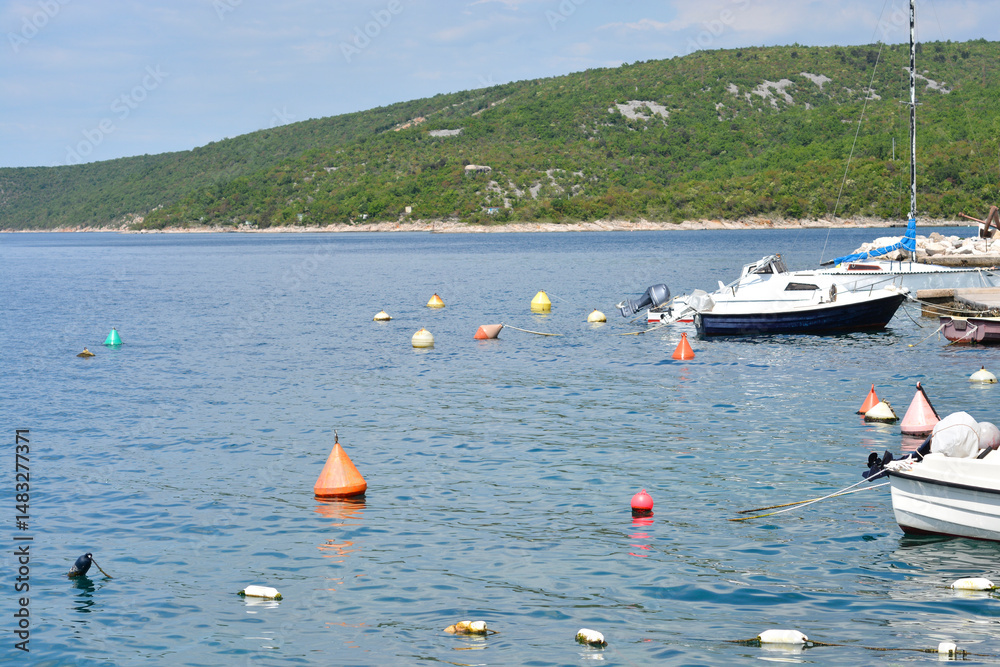 Several boats bob gently on the Adriatic Sea near Jadranovo, with buoys marking the water and a green, hilly coastline in the distance under a bright sky